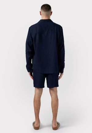 Chemise en lin bleu marine foncé à manches longues, associée à un short assorti. Coupe décontractée avec un col boutonné et des fentes sur les côtés. Sandales à lanières marron.