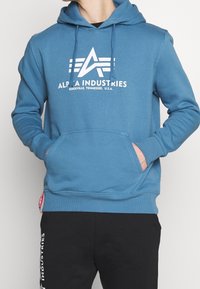 Blå bomullshuvtröja med framficka, dragsko i huvan och vit logotext. Matchas med svarta sweatpants med vit branding.