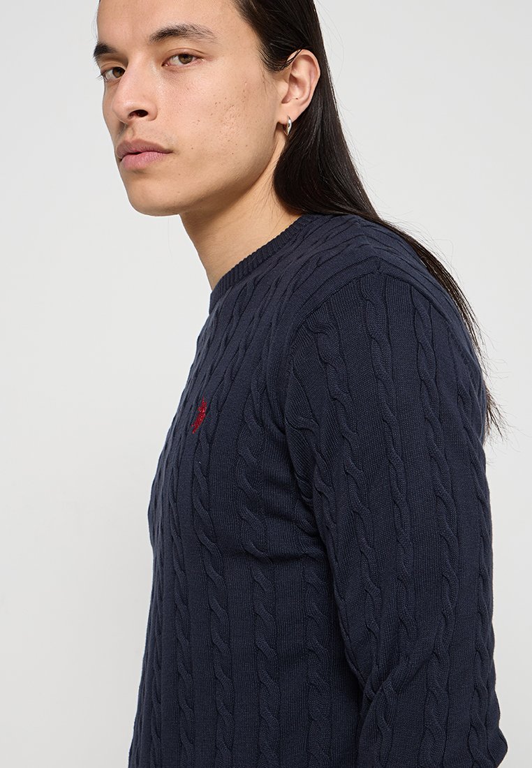 Marineblaue Kabelstrickpullover mit Rundhalsausschnitt und einem kleinen roten Logo-Detail auf der Brust. Hergestellt aus strukturiertem Strickmaterial.