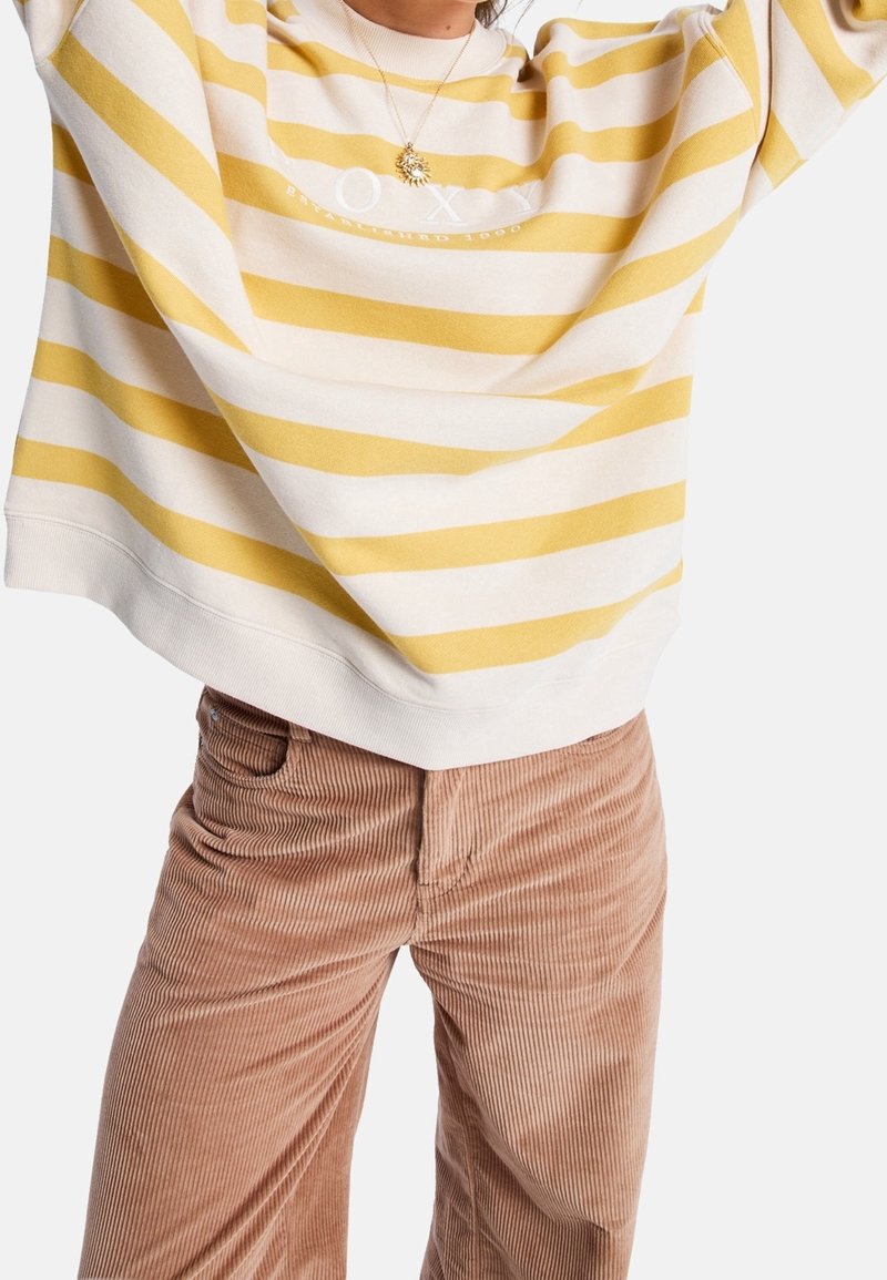 Personne portant un sweat-shirt à rayures jaunes et blanches et un pantalon en velours côtelé beige, avec un collier pendentif en forme de soleil.
