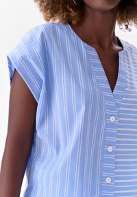 Jasnoniebieska koszula w białe pionowe paski, z dekoltem w serek, małymi guzikami z przodu i krótkimi rękawami. Gładka tekstura.
