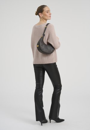 Vrouw met haar in een knot, gekleed in een beige trui, zwarte leren broek, zwarte hakken, en draagt een zwarte schoudertas in de vorm van een halve maan.