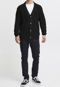 Černý pletený kardigan s výstřihem do V, s pěti knoflíky vpředu. Kombinován s tmavými úzkými kalhotami a černými teniskami.