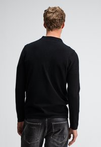 Schwarzer Pullover mit langen Ärmeln, glatter Textur, normalem Kragen und taillierter Passform, kombiniert mit dunklen Jeans aus Denim mit gesteppten Taschen.