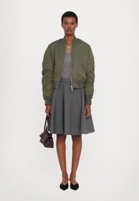 Olive green bomber jacket made of smooth material, paired with a gray sleeveless top and a flared gray skirt. Black pointed shoes and a brown bag.