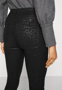 Black jeans with a shiny leopard print pattern, high waistband, and standard back pockets. Fabric has a sleek, textured appearance.
