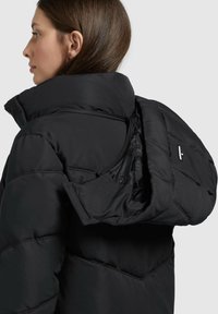 Svart puffjacka med hög krage, dragkedjestängning och en intern ficka. Den distinkta sömmen ger textur. Materialet ser slätt ut.