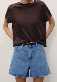 Personne portant une chemise à manches courtes marron foncé rentrée dans un short en jean bleu taille haute avec une ceinture noire, mains derrière le dos.