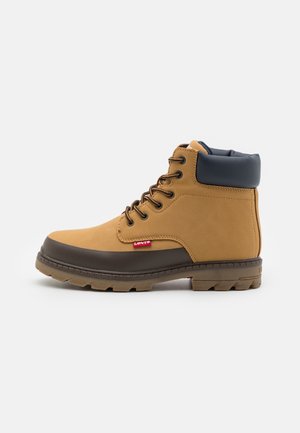 Botte en cuir marron clair avec un bracelet bleu marine, une semelle en caoutchouc brun foncé et une étiquette de logo rouge. Comprend des œillets métalliques et un design à lacets robuste.