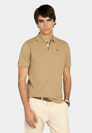 Hombre joven con una camiseta polo color beige con un logo bordado, pantalones beige y un reloj de pulsera, de pie contra un fondo claro y liso.