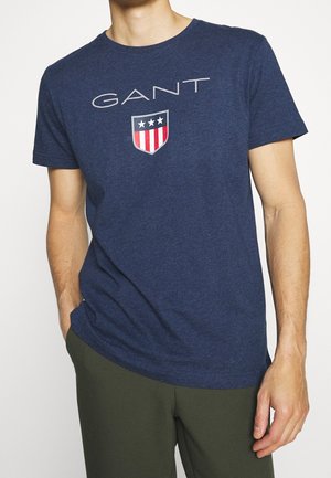 Navy blue cotton t-shirt featuring the word "GANT" and a shield logo with red and white stripes and three stars printed on the front.