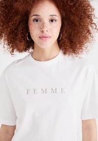 Bílý bavlněný tričko s nápisem "FEMME" v béžovém tisku. Krátké rukávy, kulatý výstřih, měkká textilie, jednoduchý design.