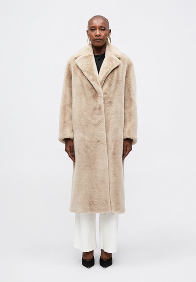 Cappotto in pelliccia sintetica beige con ampio colletto, maniche lunghe e taglio dritto, abbinato a pantaloni bianchi e scarpe con tacco nere.