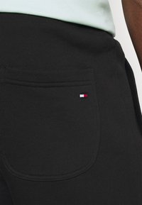Pantalones joggers negros con un acabado texturizado, que cuentan con un bolsillo trasero y un pequeño acento de logotipo en rojo, blanco y azul en el bolsillo.