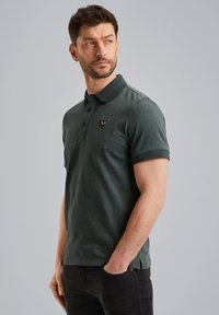Mørkegrøn polo skjorte med et tekstureret mønster, krave og tre knapper. Har et lille guldlogo på venstre bryst.