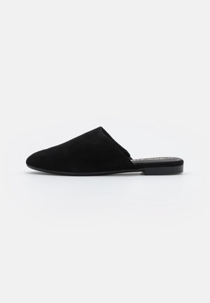 Sort ruskind slip-on mule med spids tå og flad sål, vist i profil på en ensfarvet hvid baggrund.
