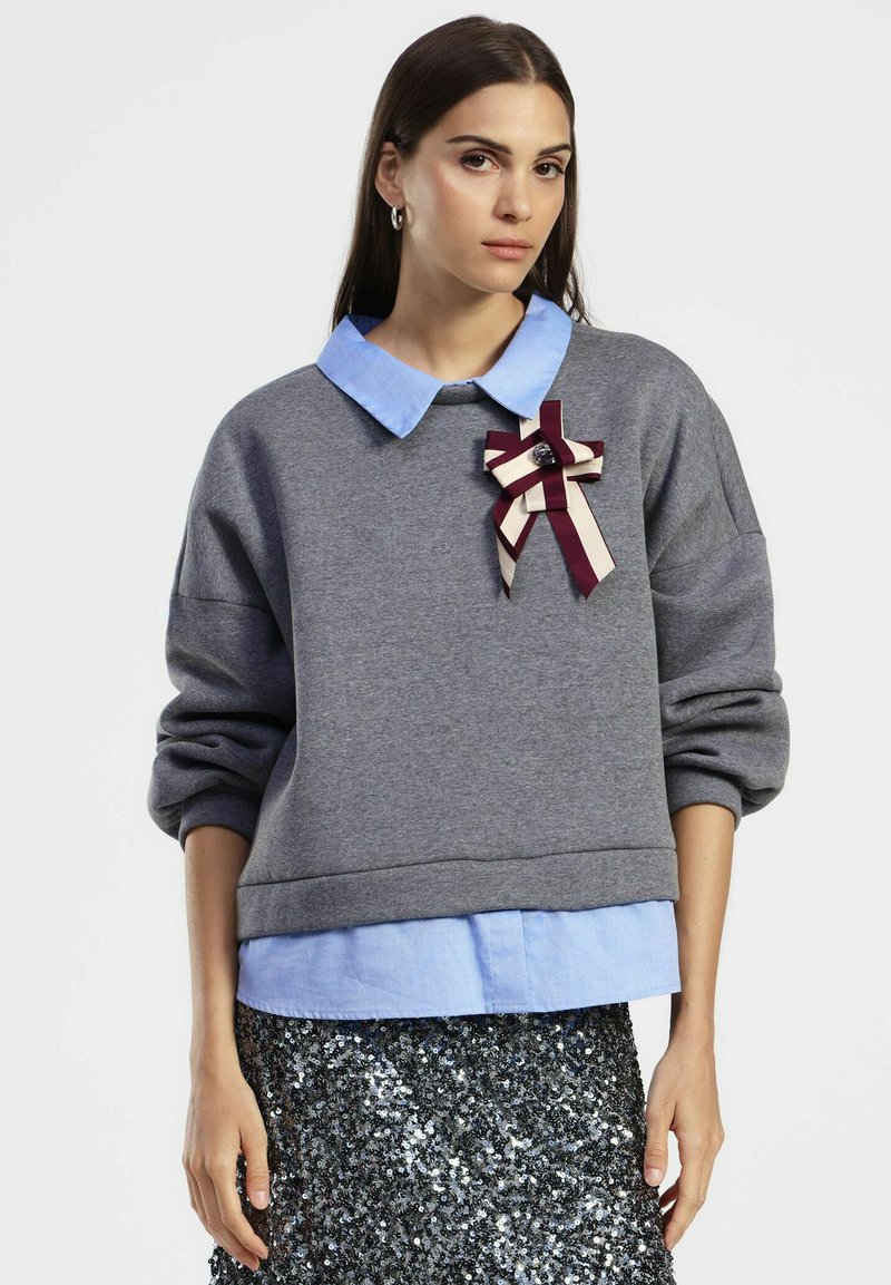 Donna che indossa una camicia blu con colletto sotto un felpa grigia con una spilla a fiocco a righe, abbinata a una gonna nera e argento con paillettes.