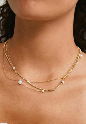 Schichtweise goldene Halskette mit Perlen und klaren Kristallen, mit unterschiedlichen Kettenlängen und -texturen, die auf einem nackten Schlüsselbein ruht.