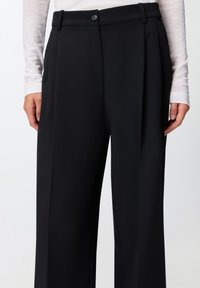 Zwarte, wijde broek in lichtgewicht stof met voorplooien, een knoopsluiting en riemlussen. Soepele textuur, nauwsluitende pasvorm.