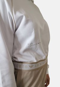 Valkoinen collegepaita beigeilla yksityiskohdilla, jossa on EA7 Emporio Armanin logo. Elastinen vyötärö matching beige -värissä brändityksillä.