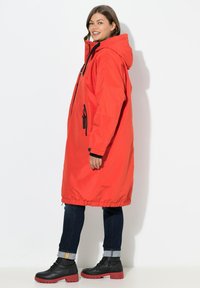 Parka rouge imperméable avec capuche, dotée d'une fermeture éclair noire et de poches. Portée sur un jean foncé et des bottes noires avec des semelles rouges.