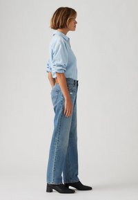 Chemise bleu clair à manches retroussées, associée à un jean bleu délavé taille haute et des bottes noires à talons, vue de côté.