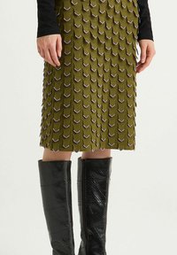Jupe texturée vert olive avec un design en couches et des accents violets, associée à des bottes montantes en cuir verni noir avec une fermeture éclair.