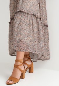 Robe maxi florale avec un ourlet superposé et volanté dans des tons de rose et de bleu atténués, associée à des sandales à talons blocs beiges avec des brides entrecroisées.