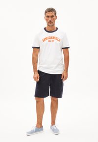 Witte t-shirt met marineblauwe accenten en oranje tekst, gecombineerd met marineblauwe shorts en lichtblauwe sneakers. Casual ontwerp, katoenen materiaal.