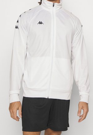 Homme portant une veste de sport blanche zippée avec des logos noirs sur la poitrine et les manches, associée à un short de sport noir.