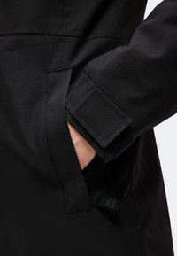 Main insérée dans la poche latérale d'une veste noire avec un poignet réglable en Velcro sur la manche.