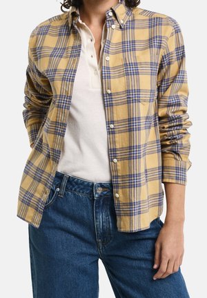 Persoon draagt blauwe spijkerbroek, wit blouse met knopen en een geel-blauw geruit blouse met opgerolde mouwen.