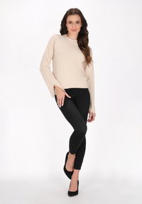 Pulover bej tricotat cu mâneci lungi și evazate, asortat cu colanți negri și tocuri înalte negre. Modelul pozează cu o mână pe șold.