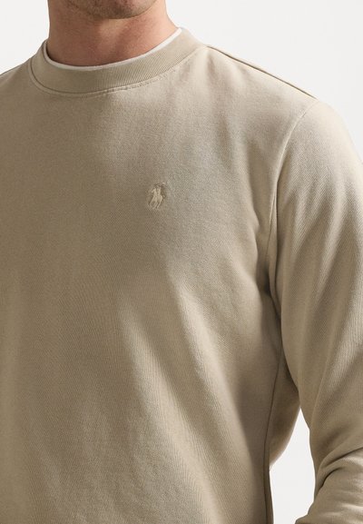 Beige hosszú ujjú pulóver, kerek nyakkivágással, texturált anyagból, a mellrészen egy finom hímzett logóval.