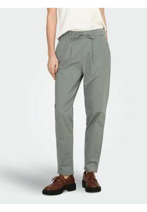 Personne portant un pantalon gris clair fuselé avec une taille à cordon de serrage et des chaussures en cuir marron à lacets, debout devant un fond uni.