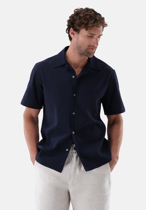 Mann mit lockigem Haar, der ein marineblaues kurzärmeliges Hemd und hellbeige Hose trägt, Hände in den Taschen, schaut nach unten links.