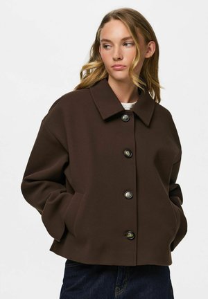 Jeune femme portant un manteau marron foncé boutonné, les mains dans les poches, regardant vers sa gauche avec des cheveux blonds ondulés et lâches.
