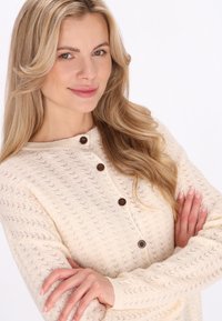 Roomkleurig gebreid vest met een gestructureerd patroon, een ronde halslijn en drie bruine knopen. Lange mouwen zorgen voor een comfortabele pasvorm.