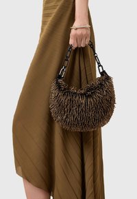 Hnedá texturovaná kabelka s okrúhlym tvarom a vystupujúcim detailom, držená čiernym reťazovým uškom, nosená s rebrovanými hnedými šatami.