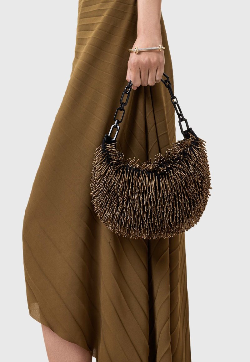 Hnedá texturovaná kabelka s okrúhlym tvarom a vystupujúcim detailom, držená čiernym reťazovým uškom, nosená s rebrovanými hnedými šatami.