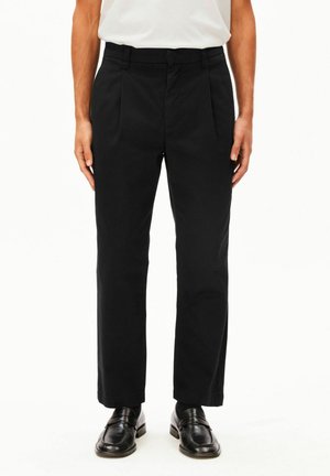 Homme portant un pantalon de costume noir, des chaussures habillées noires et une chemise blanche, debout les bras détendus le long du corps devant un fond uni.