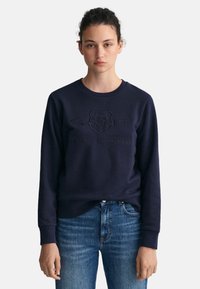 Felpa navy realizzata in tessuto morbido, caratterizzata da un logo in rilievo con un design a scudo. Polsini e orlo a coste con scollo tondo.