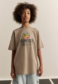 Camiseta de algodón beige con diseño de frutas bordado y texto "LOS ÁNGELES". Corte holgado con mangas cortas, escote redondo y textura suave.