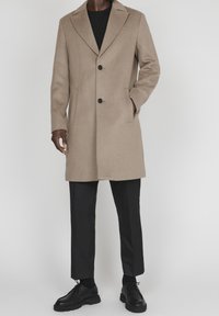 Hombre vestido con un abrigo beige hasta la rodilla sobre una camisa negra, pantalones negros y zapatos de cordones negros, de pie frente a un fondo liso.
