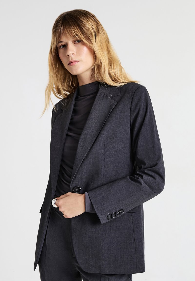 Donna con lunghi capelli biondi che indossa un blazer grigio scuro su un top nero a collo alto, in piedi su uno sfondo chiaro uniforme.