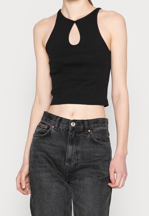 Frau trägt ein schwarzes, ärmelloses Crop-Top mit tropfenförmigem Ausschnitt und hoch taillierte, ausgewaschene schwarze Jeans, steht vor einem einfarbigen Hintergrund.