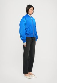 Kvinde iført en lysblå lynlås bomberjakke, grå turtleneck sweater, sorte lige jeans og hvide spidse hæle.