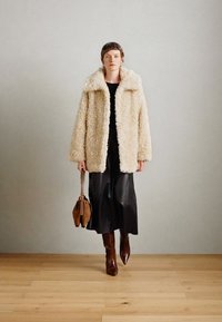 Kreemivärvi faux fur mantel, üleriie suurustega, laia kraega, kantud musta toppi ja pika musta seeliku peal. Tumedat pruuni käekotti ja saapaid täiendab välimust.