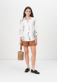 White floral-patterned shirt with 3/4 sleeves, brown suede shorts, black loafers, and a woven round bag with a strap.