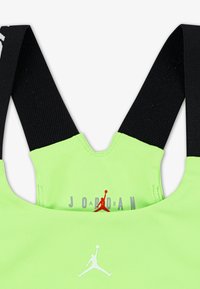 Zaļš sporta krūšturis ar melnām elastīgām siksnām. Iekļauj balto Jumpman logotipu un "JORDAN" pelēkā krāsā. Gluda tekstūra un pieguļošs dizains.